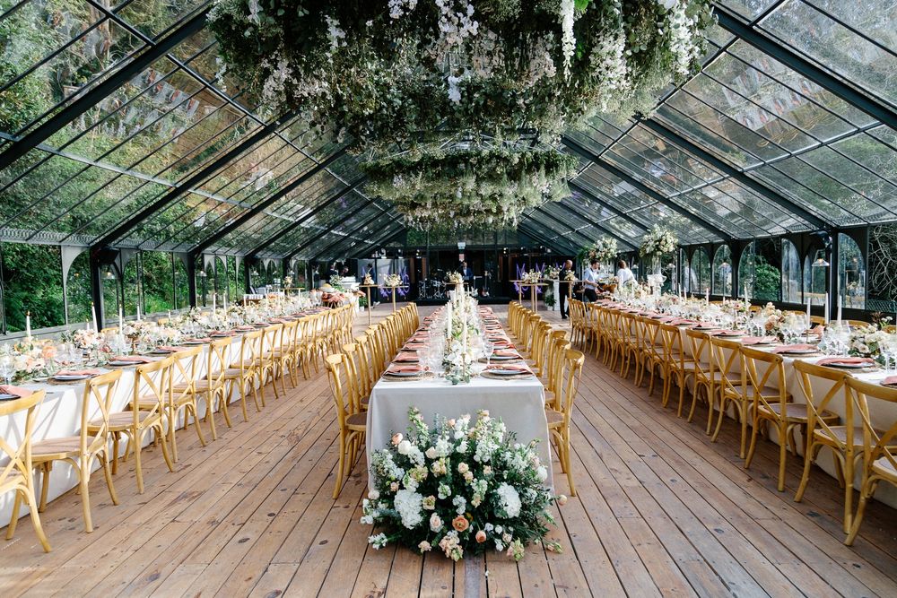 Glass greenhouse wedding reception venue with wooden chairs and white floral decorations.