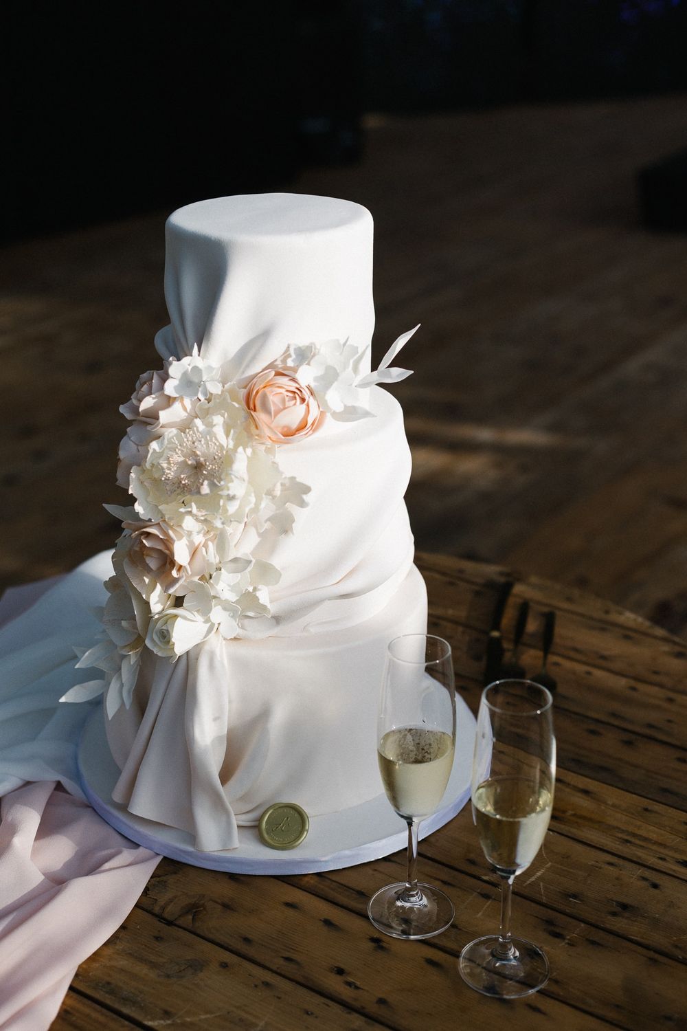 Elegant white wedding cake with delicate floral details and two champagne glasses on a rustic wooden table.