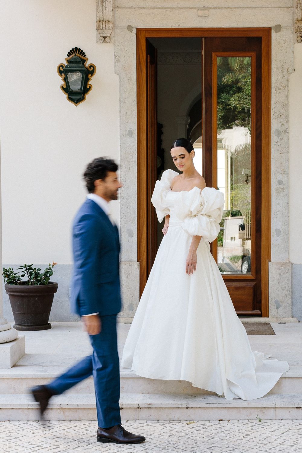 Bride in white puff sleeve gown stands in doorway as groom in blue suit walks past.