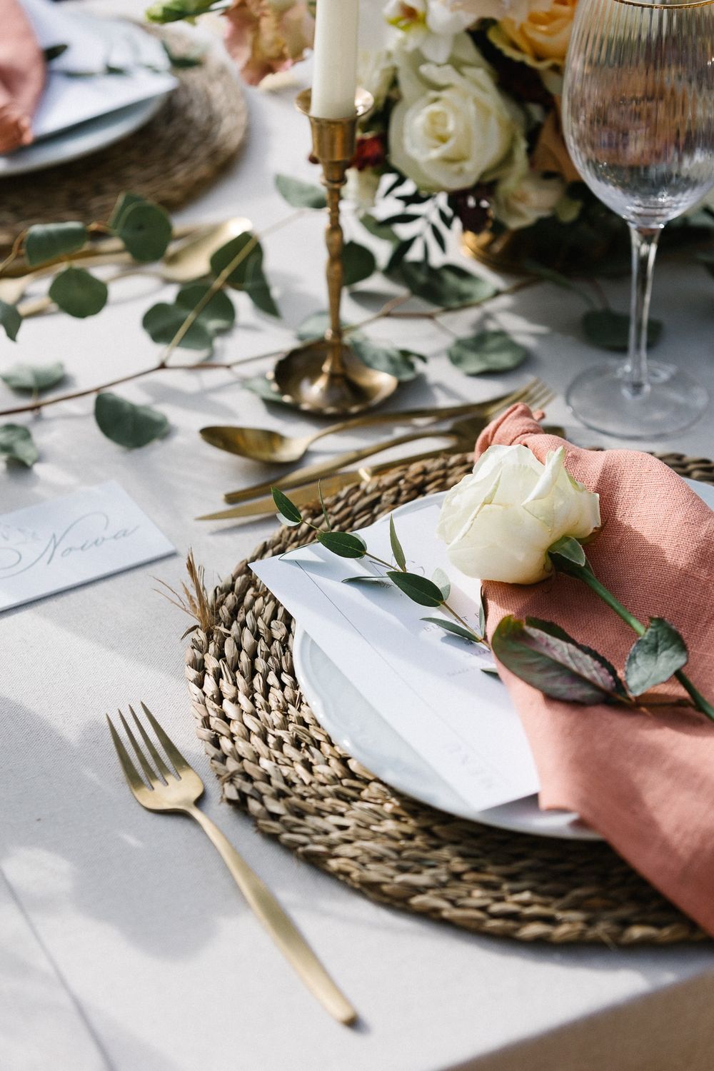 Place setting with woven charger, peach napkin, white rose and gold flatware on white tablecloth.