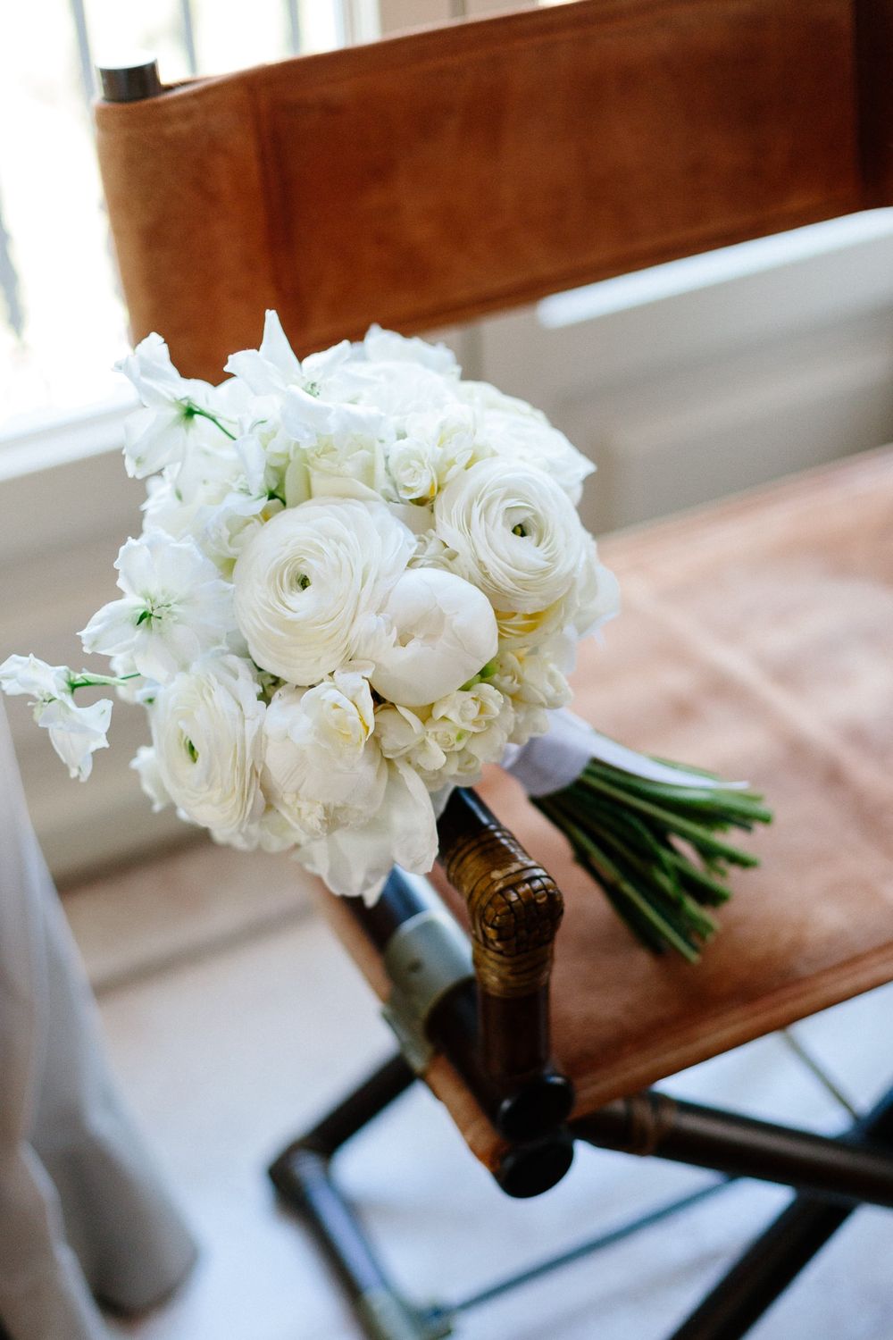 Classic white ranunculus and rose bridal bouquet resting on leather chair.