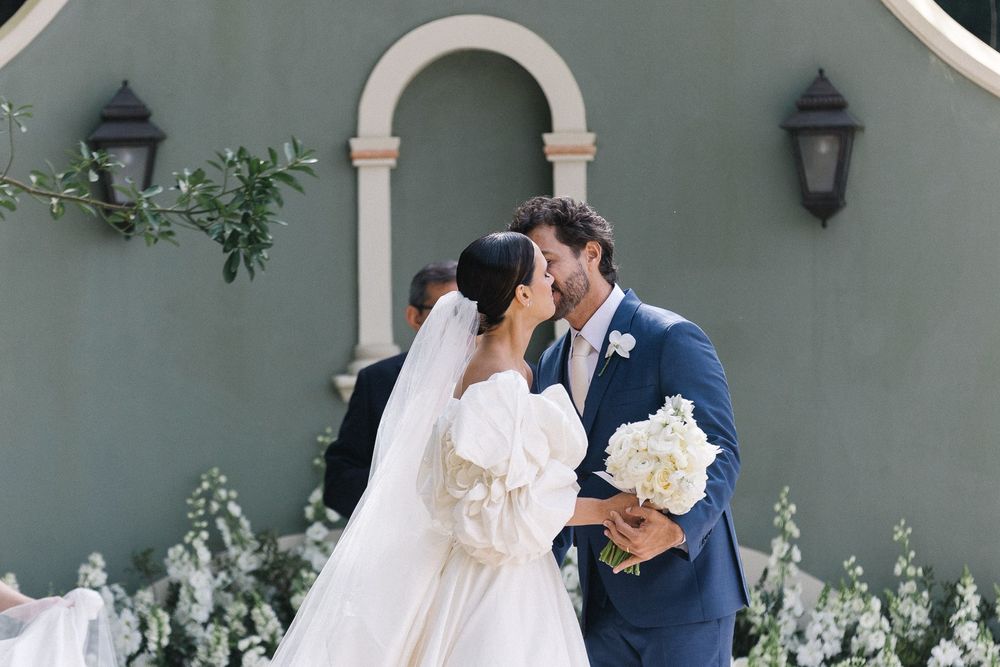 A romantic wedding moment captured in front of a grey wall with ornate architectural details and lanterns.
