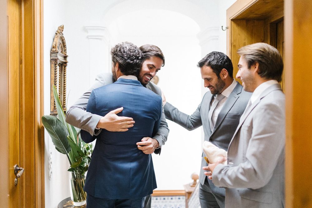 Group of formally dressed men embracing and celebrating in a bright hallway.