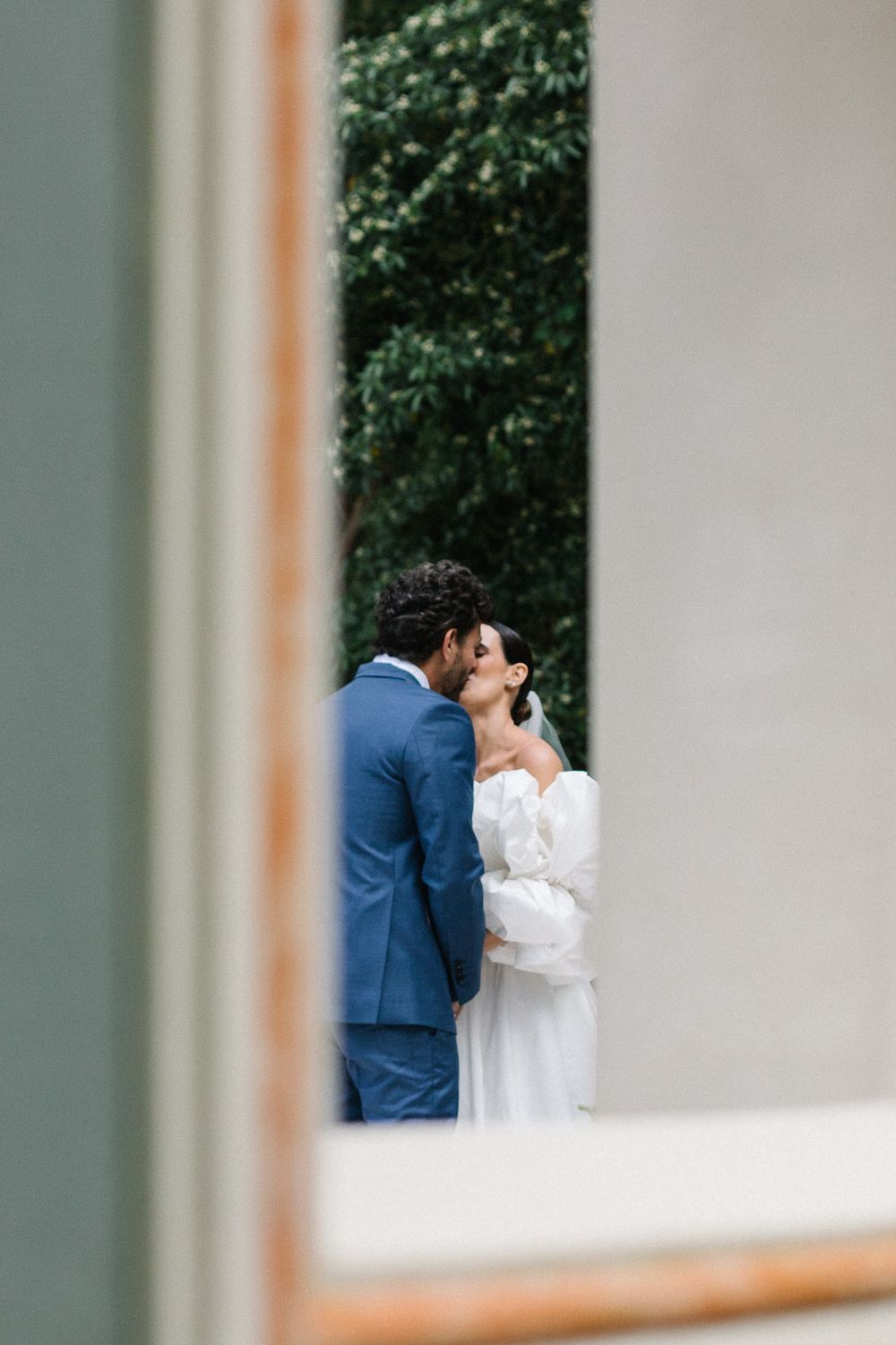 An intimate wedding moment framed through architectural elements showing couple in formal attire.