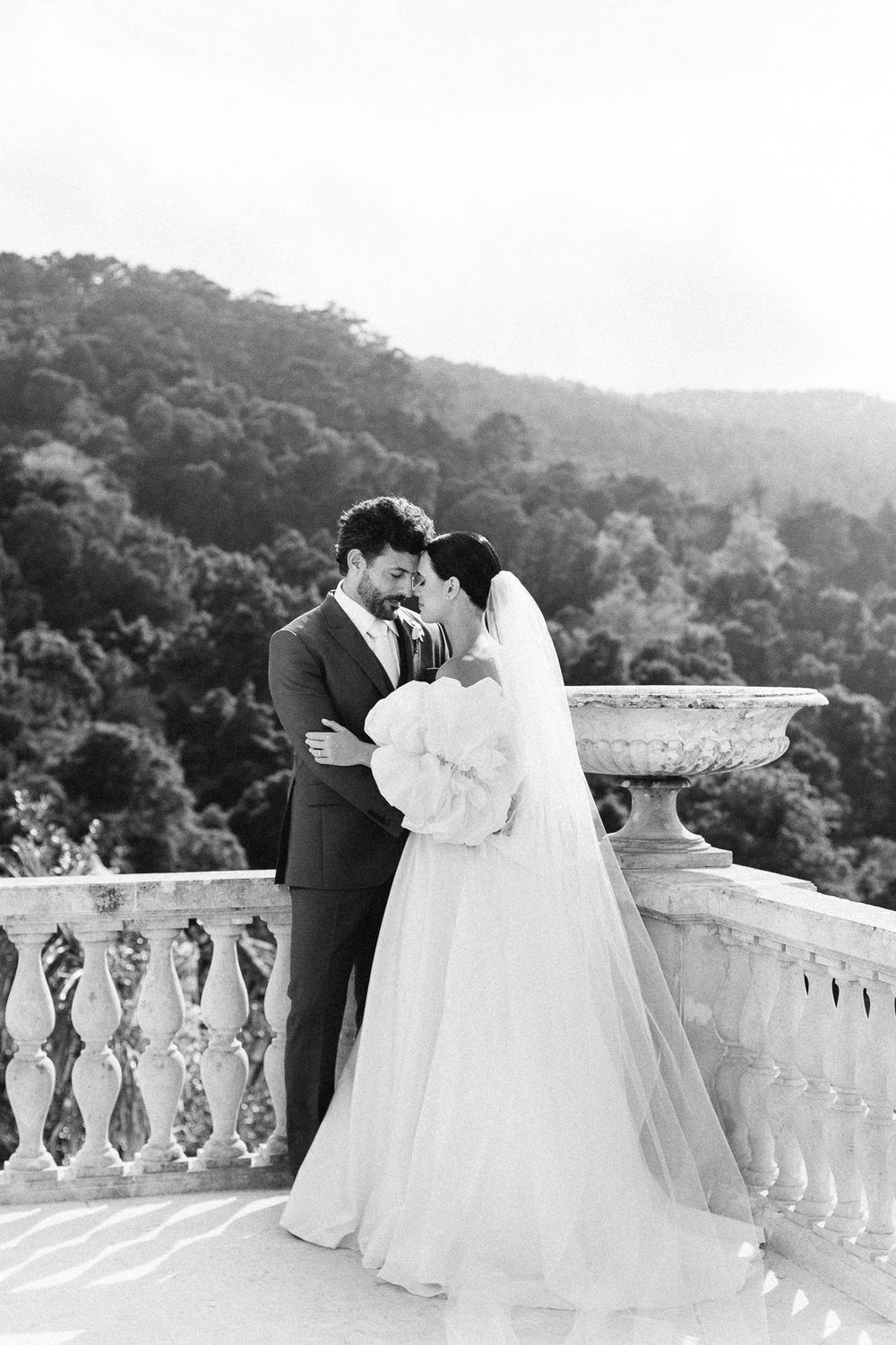 Black and white portrait of newlyweds embracing on a stone balcony with mountain views.