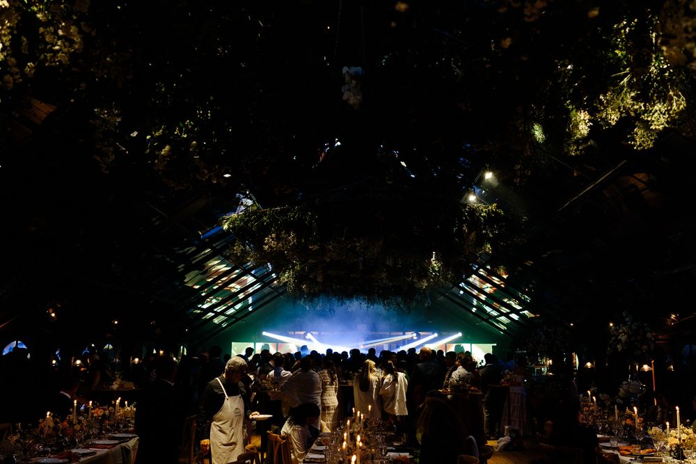 Nighttime wedding reception in illuminated greenhouse with guests on dance floor.