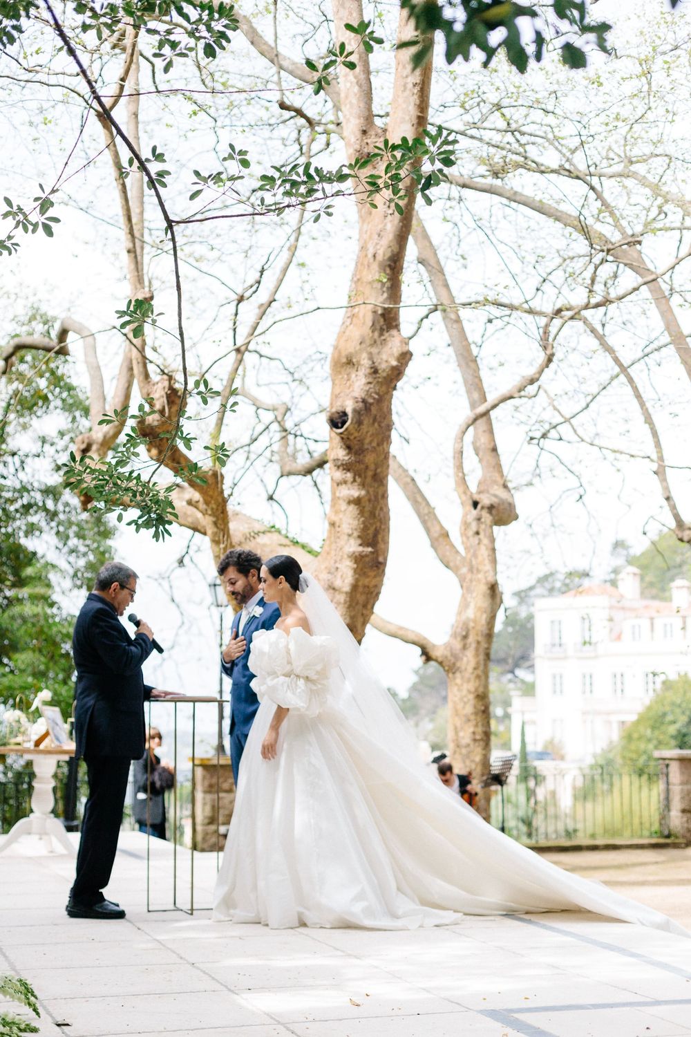 Photographer captures wedding couple under tall trees at elegant outdoor venue.