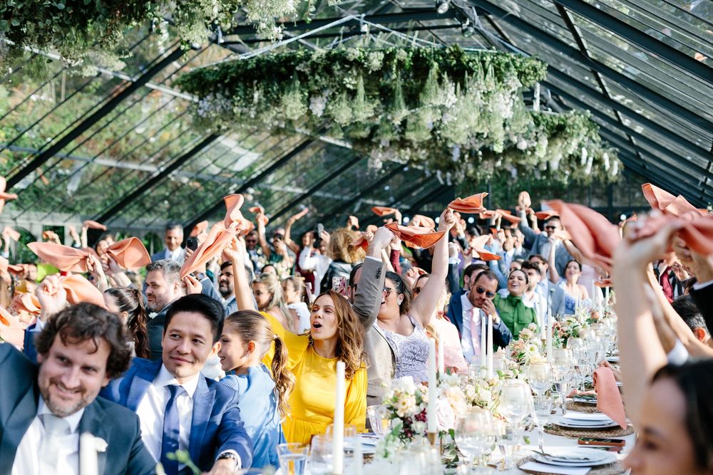 Joyful wedding guests celebrating under a greenhouse ceiling decorated with hanging flowers.