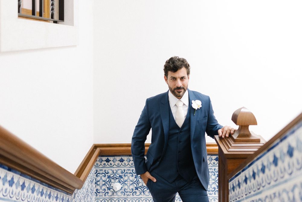 A dapper groom in a navy blue three-piece suit stands by an ornate staircase with decorative tiles.