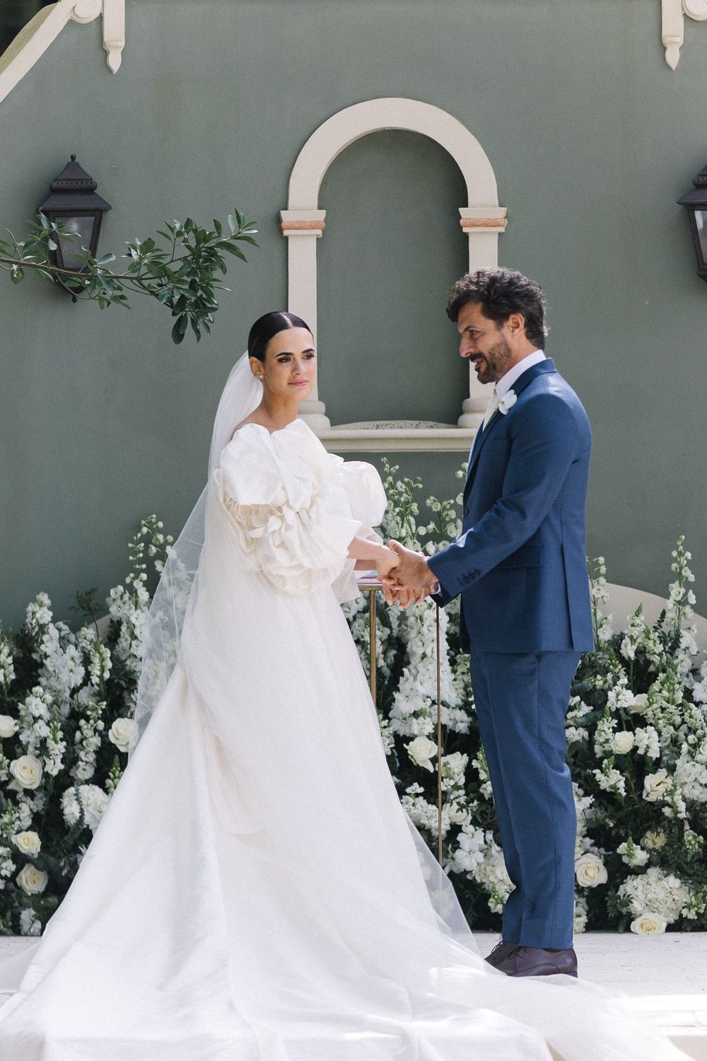 Elegant wedding couple posing against a sage green wall with white floral arch.