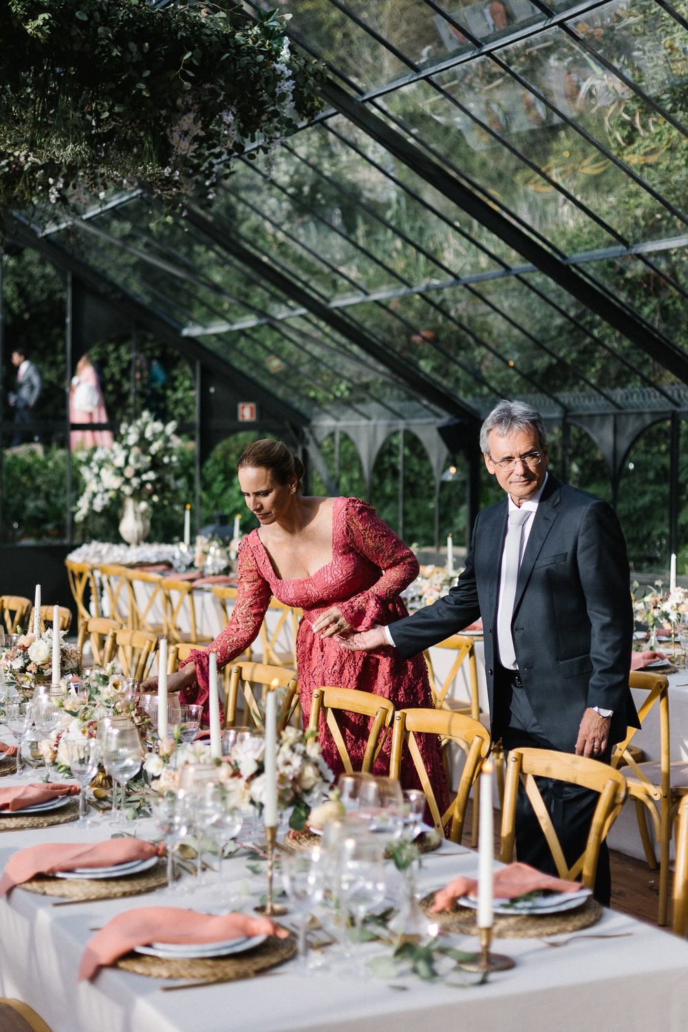 Wedding reception tables set inside elegant greenhouse with wooden chairs and floral decorations.