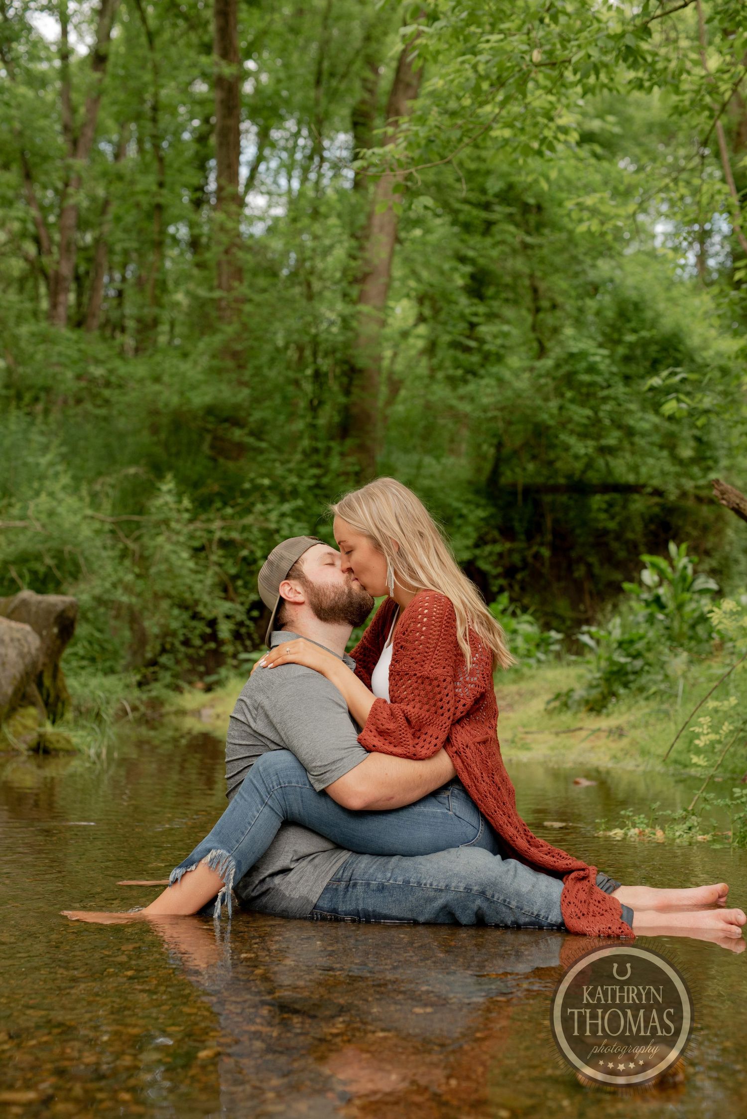 A couple shares an intimate moment while sitting in a shallow creek with forest backdrop.