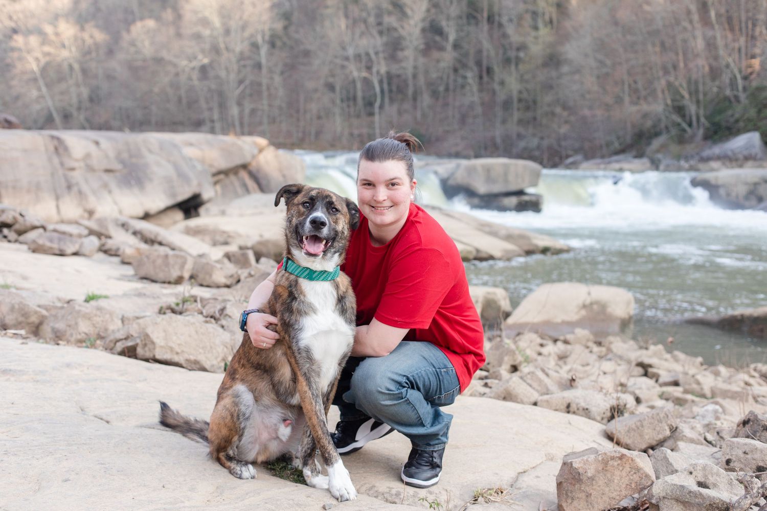 A person in red kneels beside their happy brindle dog against a waterfall backdrop.
