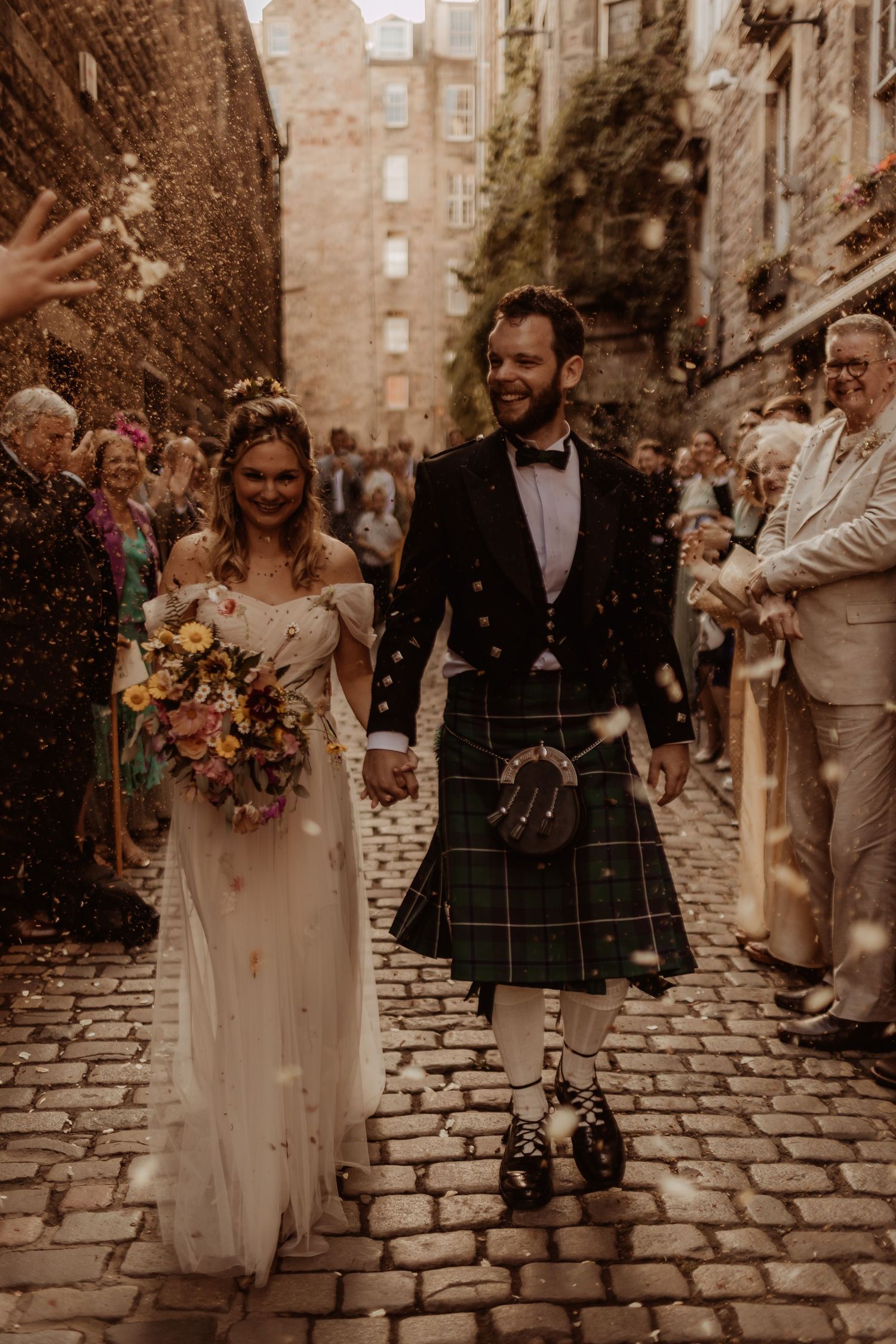 Newlyweds walk down cobblestone street while guests throw confetti, groom wearing traditional Scottish kilt.