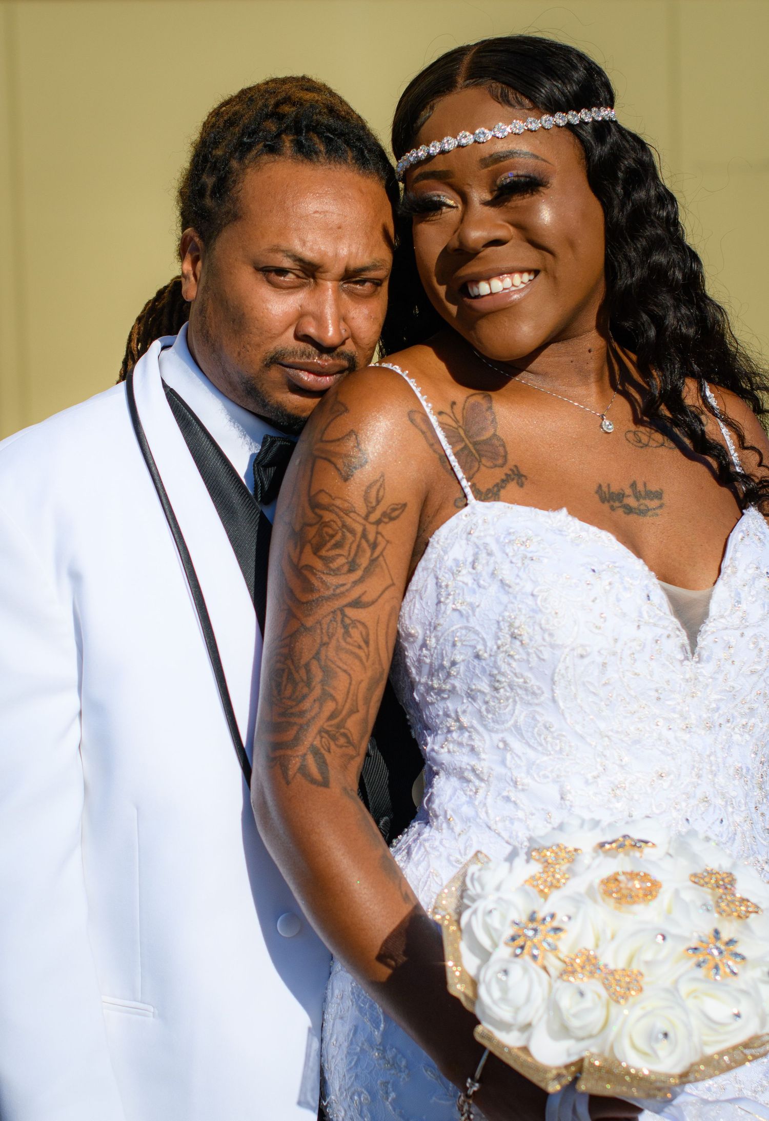A couple in wedding attire pose together, with the bride wearing a lace dress and headpiece and showing decorative arm tattoos.