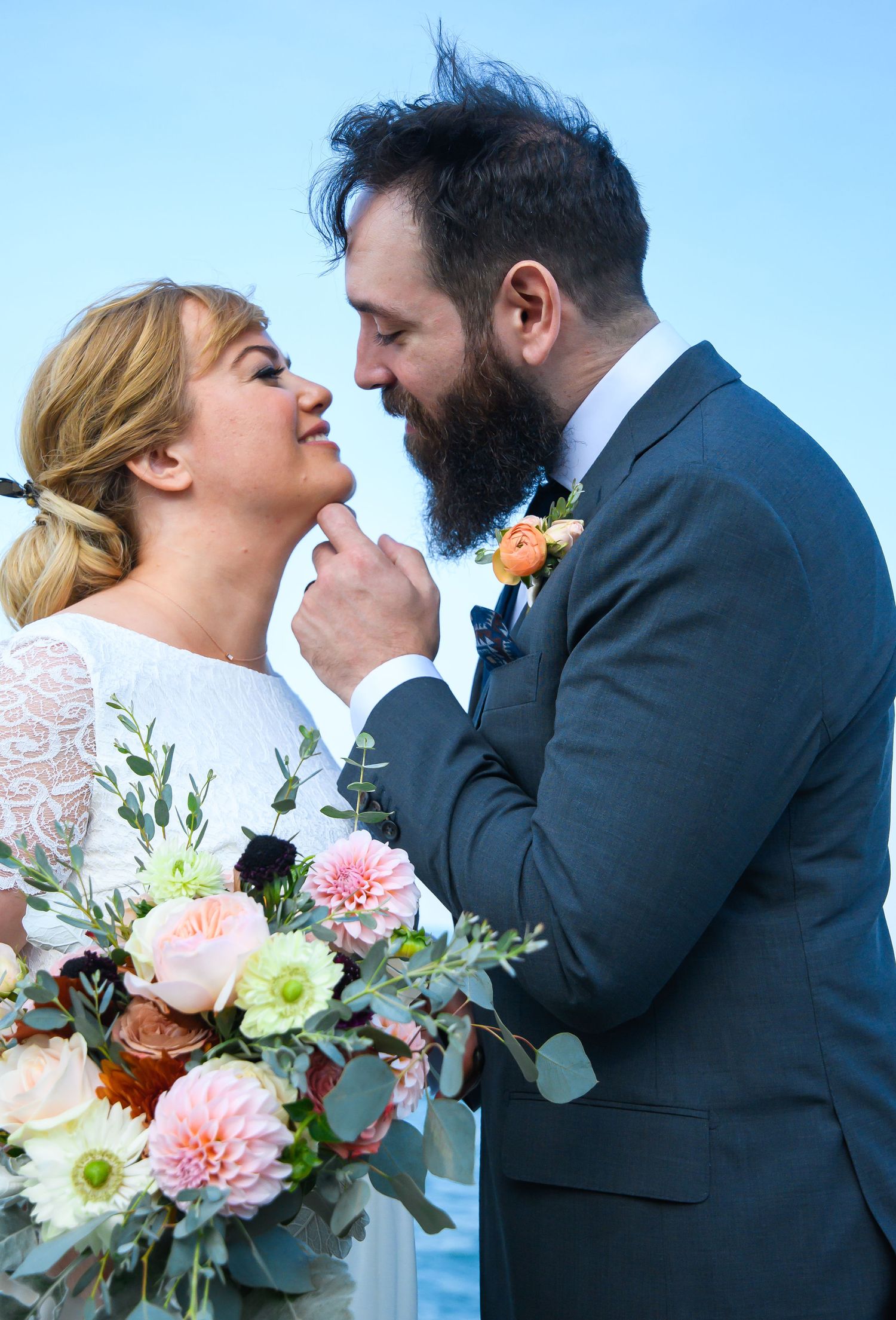 Romantic wedding couple sharing intimate moments with floral bouquet against blue sky.