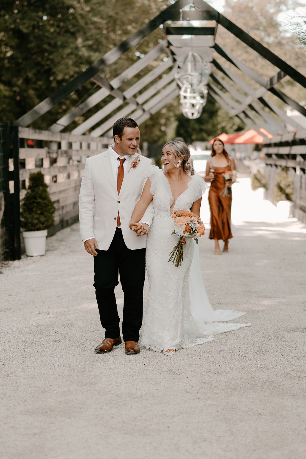 A couple walks together on a gravel path wearing elegant wedding attire.
