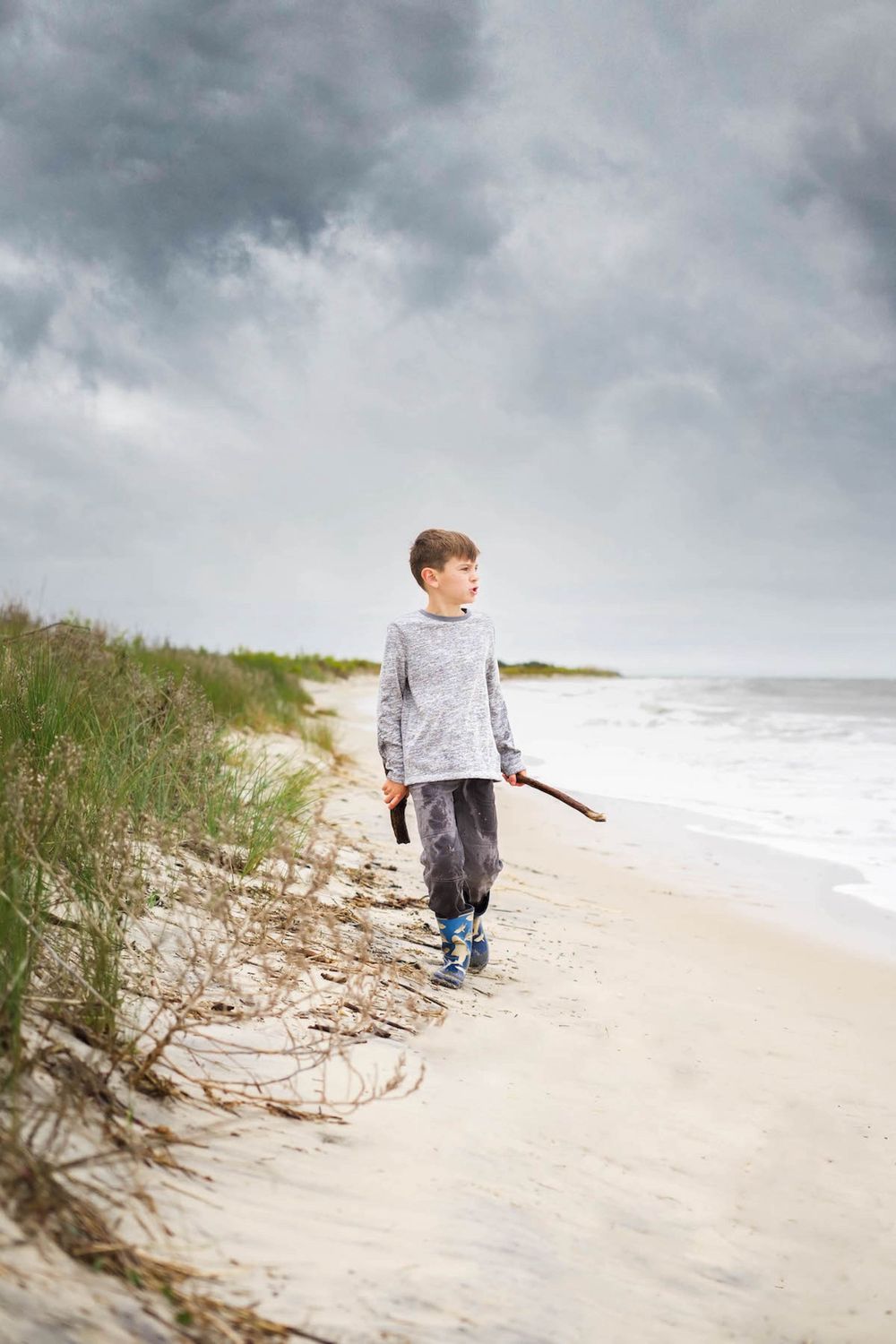 little boy walking on a beach holding a stick with a cloudy sky