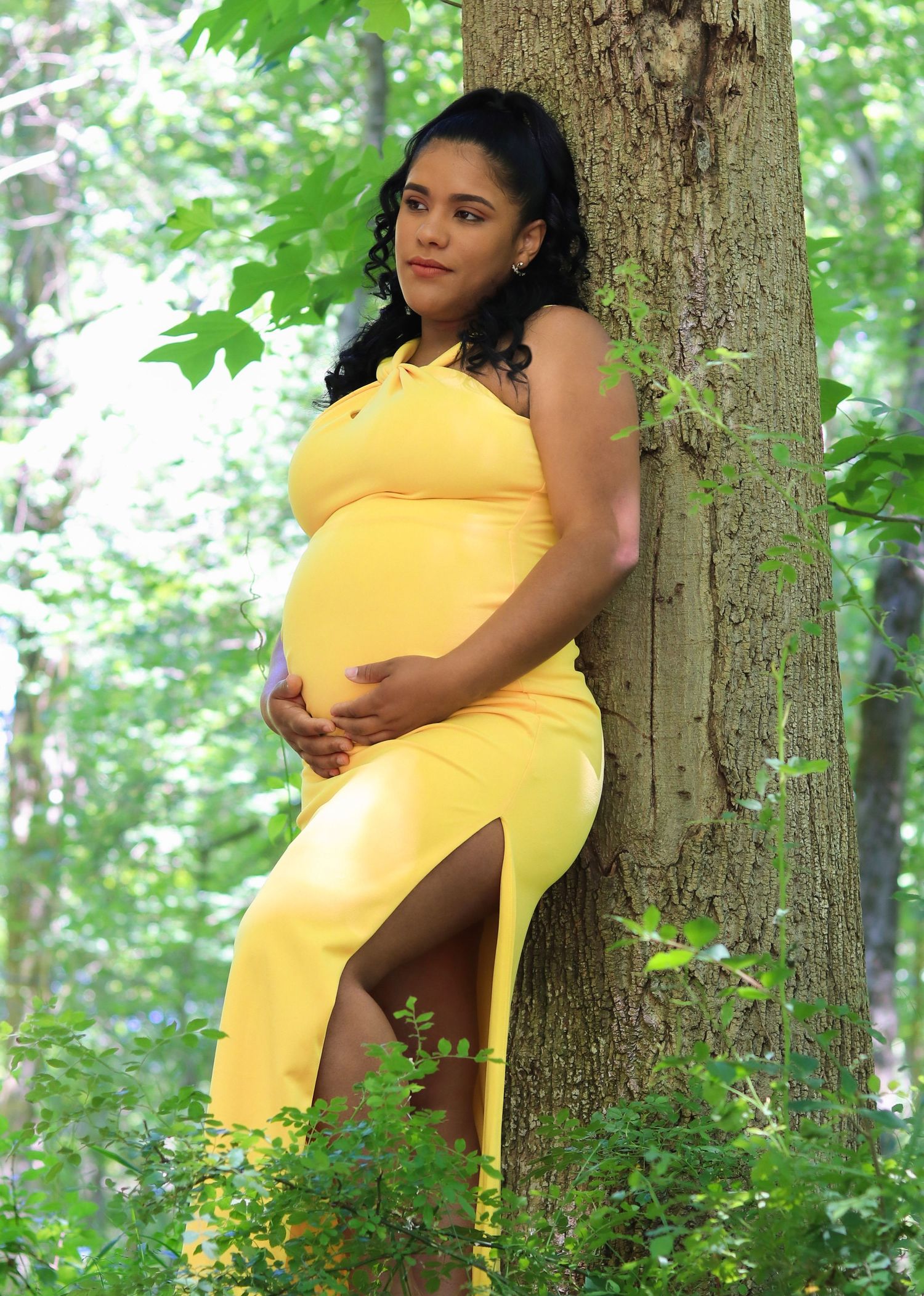 A person in a bright yellow dress poses against a tree trunk in a wooded outdoor setting during a maternity photoshoot.