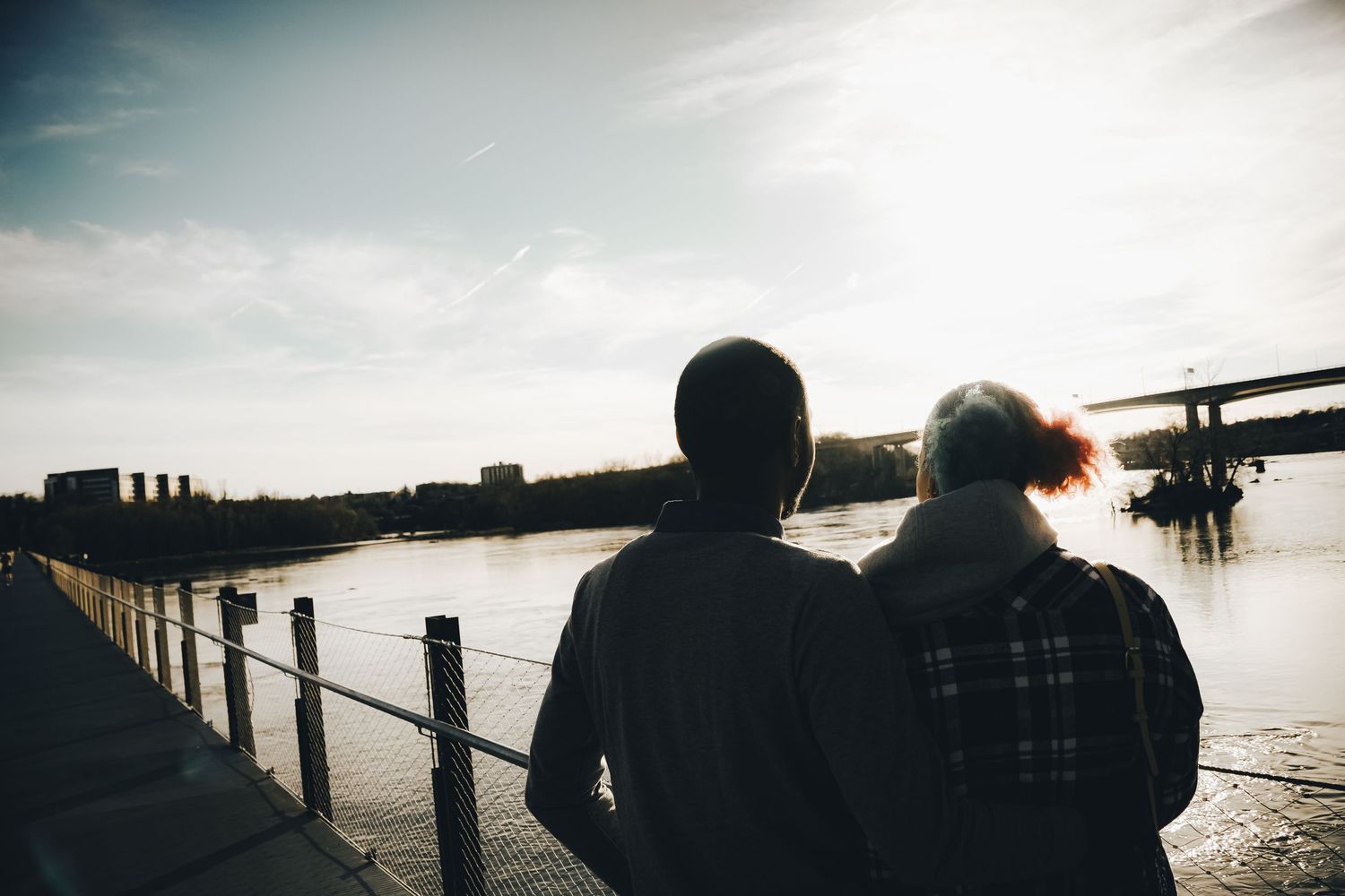 Silhouettes gaze at sunset over waterfront with railing and bridge in distance.