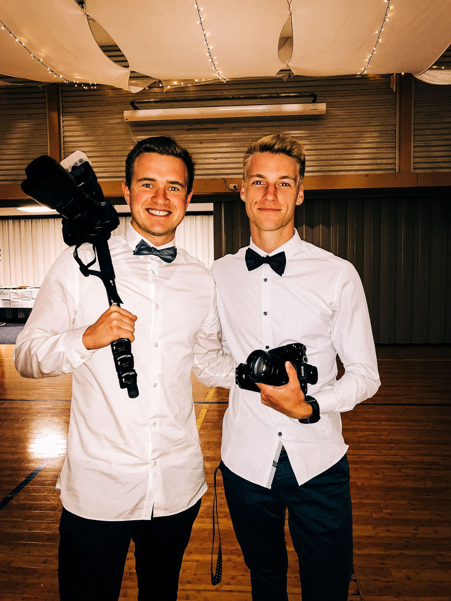 Two photographers in formal white shirts and bow ties holding professional camera equipment in an indoor venue.