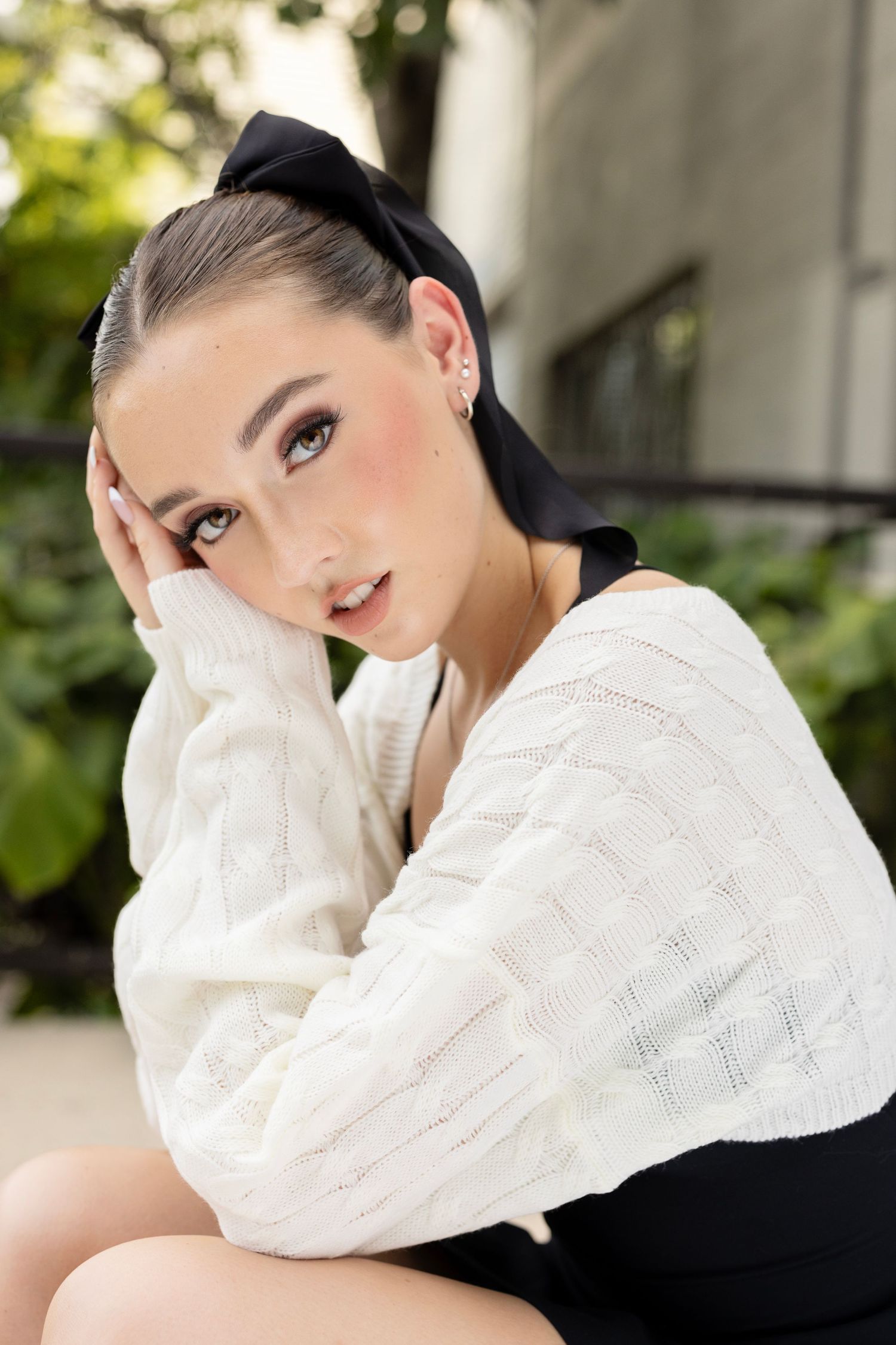 Portrait against greenery featuring a classic beauty look with a black bow and white knit sweater.