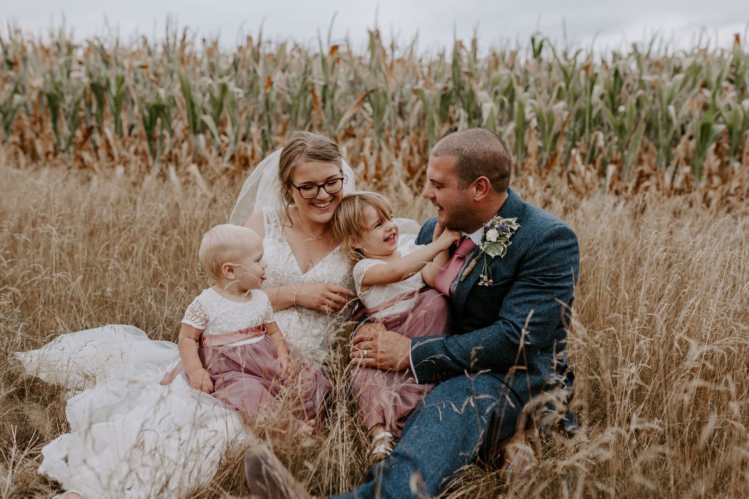 a bride and groom are sat down in long grass with their two daughters who are bridesmaids. they are all laughing with each other.