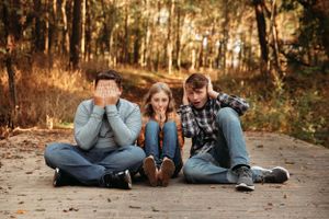 Family of three sitting on a woodland path with parents covering their eyes during a fun photo session with L. Ballenger Photography.