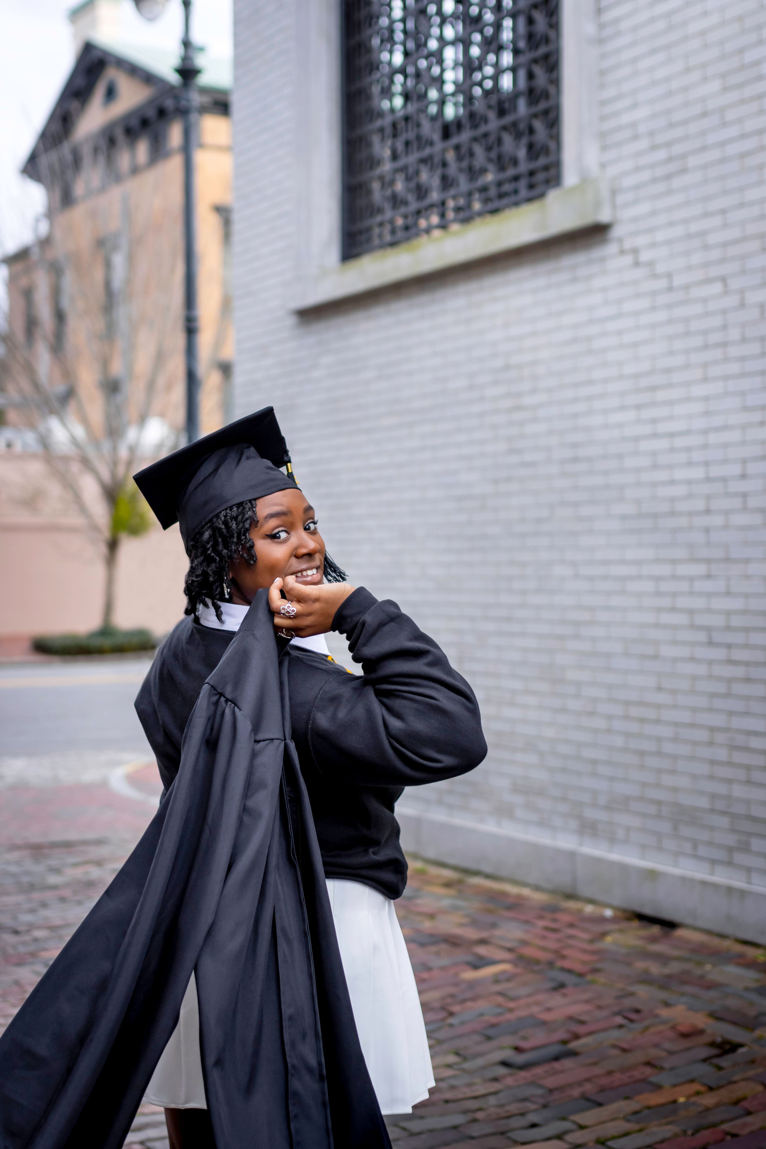 Playful graduation photo of graduate in cap and gown posing against white brick building.