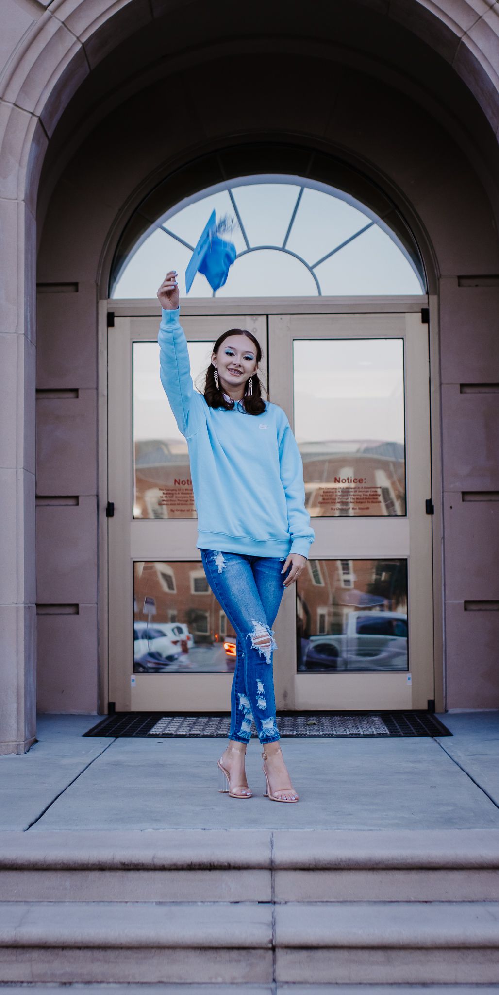 Person in light blue sweater poses joyfully on steps of building with arched entrance.