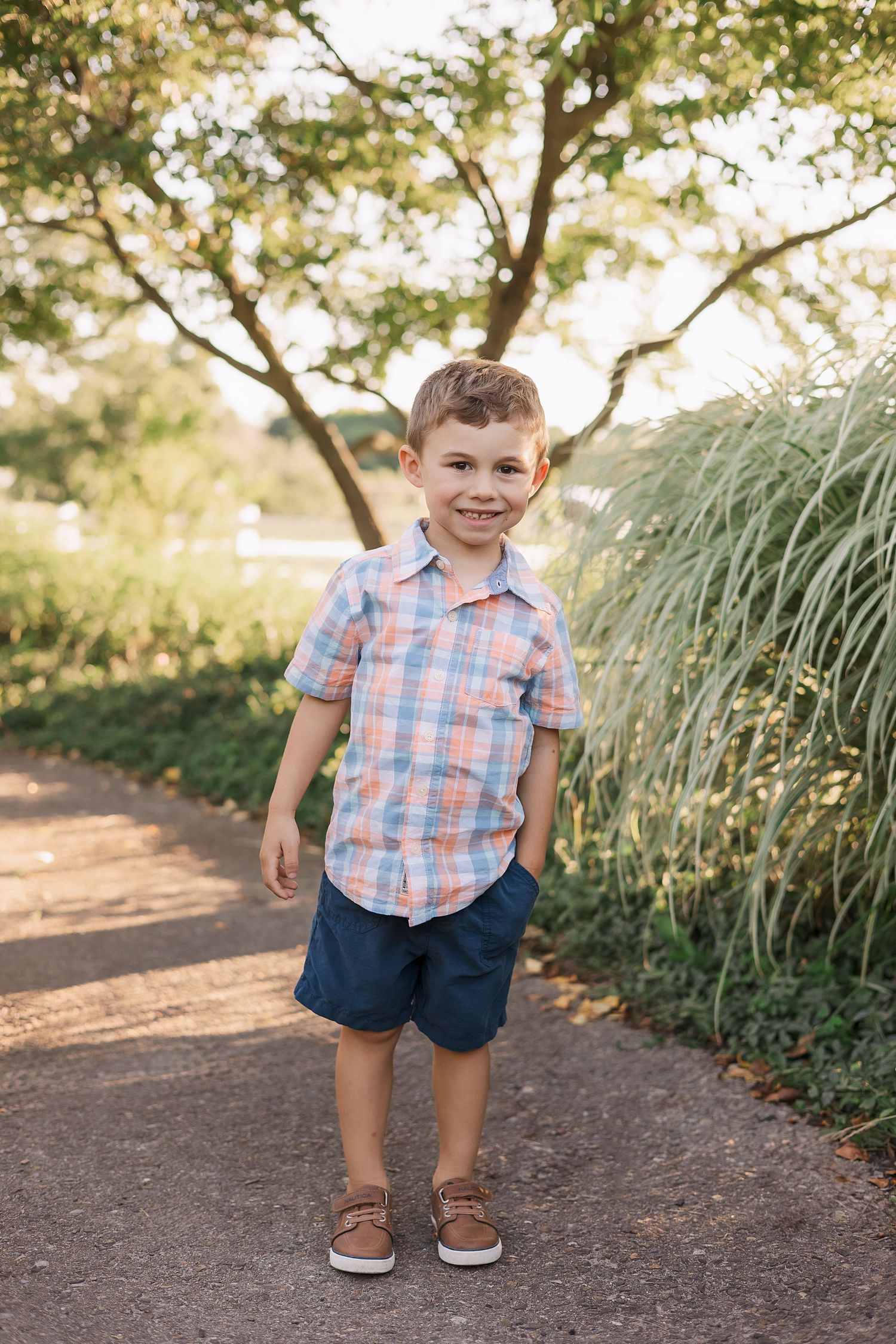 Child in plaid shirt and shorts stands on garden path near ornamental grasses. Novi Northville Portrait Photographer