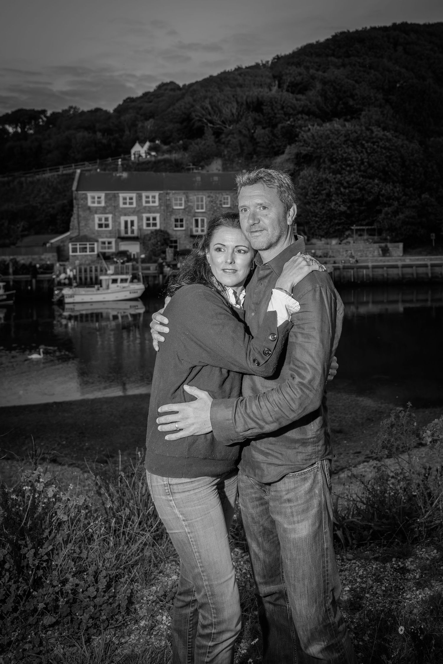 Romantic couple embraces at dusk by a waterfront with historic buildings and hills in the background.