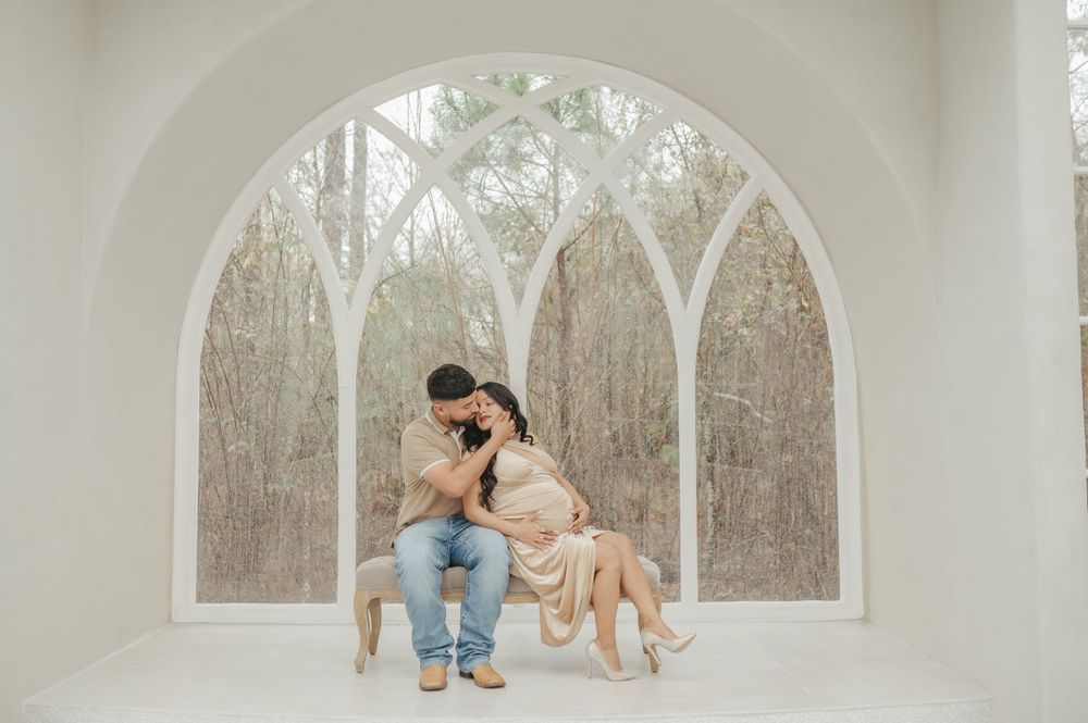 Intimate maternity photo in white room with arched window and winter scene outside.
