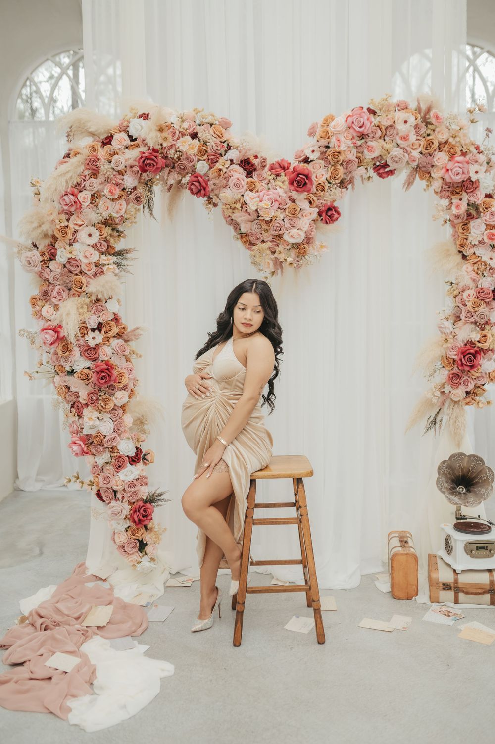 Glamorous maternity photo with floral arch and vintage props in studio setting.