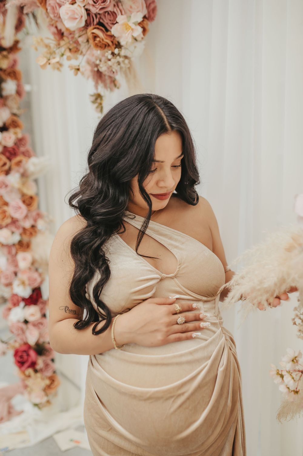 Expectant mother in champagne dress poses against floral backdrop during maternity shoot.