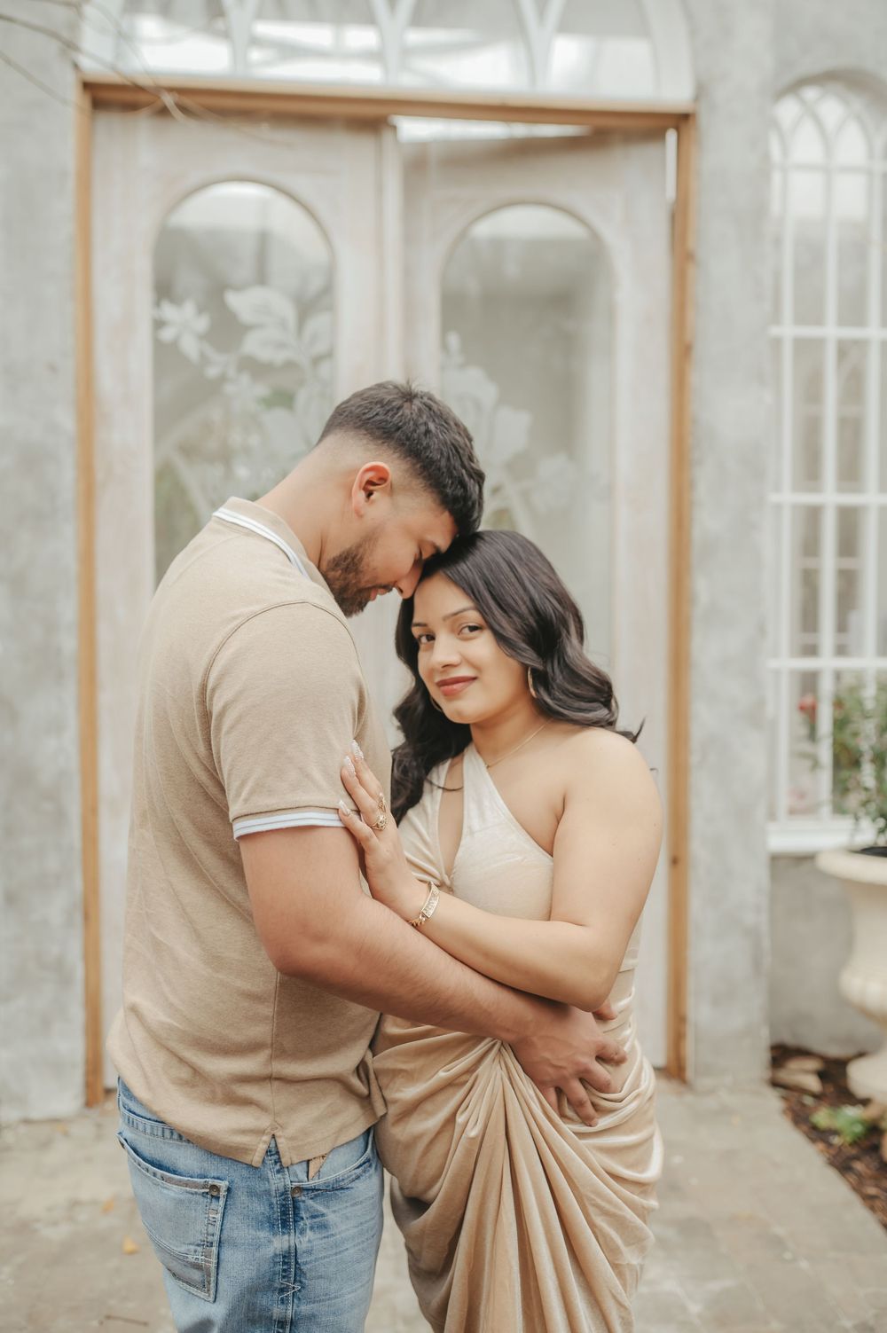 Romantic couple embraces in an elegant conservatory with arched windows and natural light.