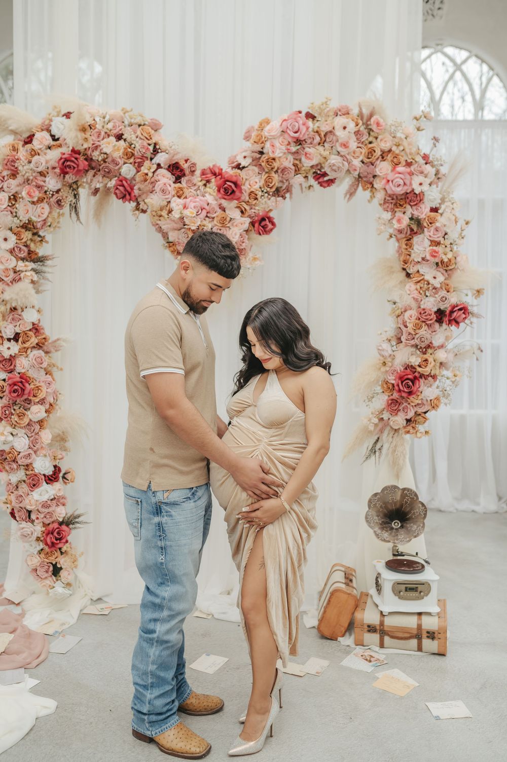 Maternity photoshoot with floral arch featuring pink and peach roses against white backdrop.