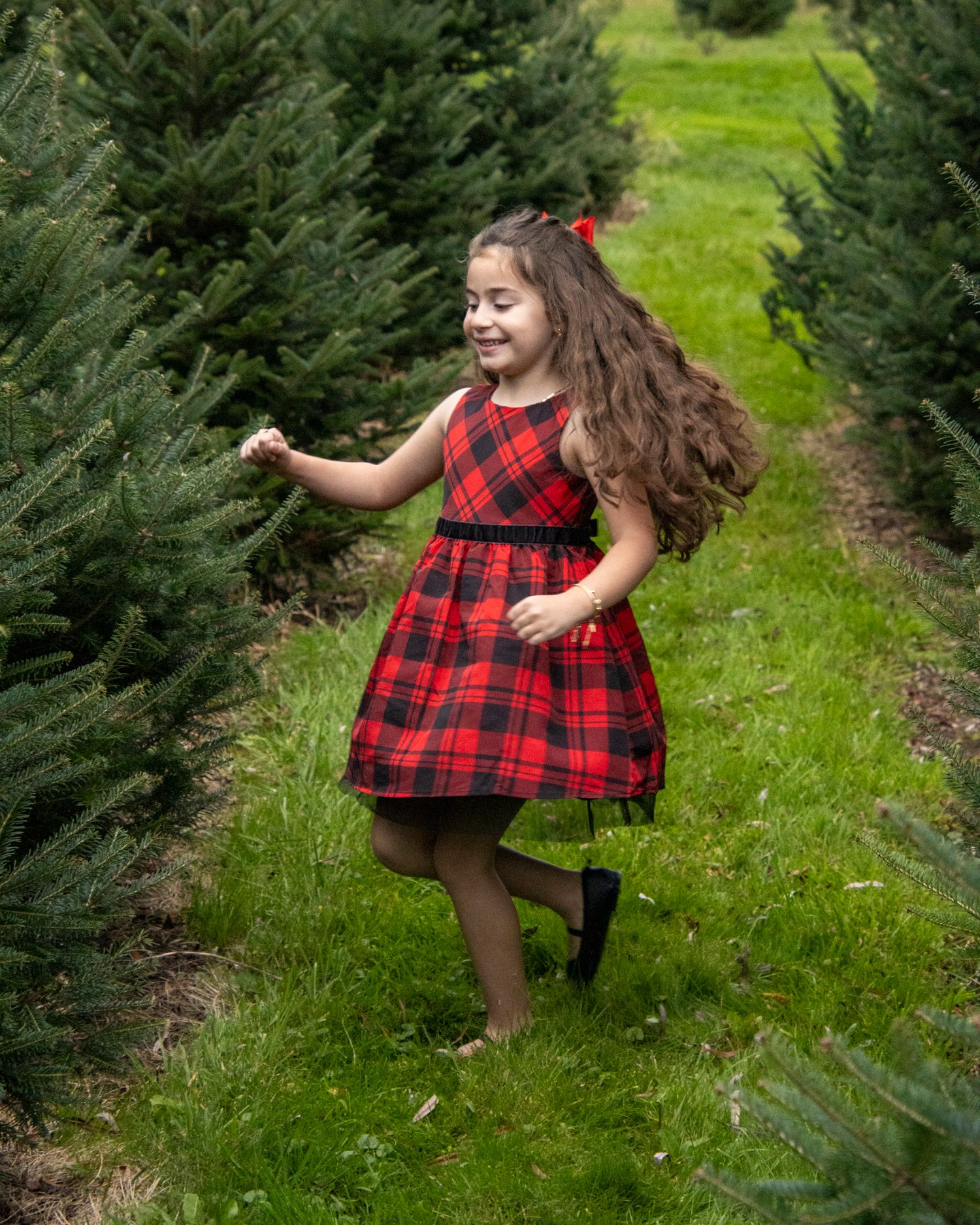 Young girl in red plaid dress dances joyfully between Christmas trees at a tree farm.