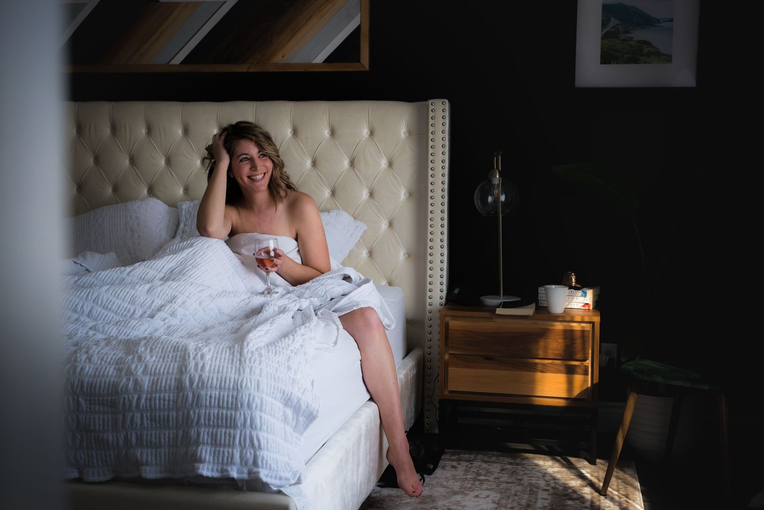 Person wearing white sits on an upholstered bed in a moody boudoir setting.