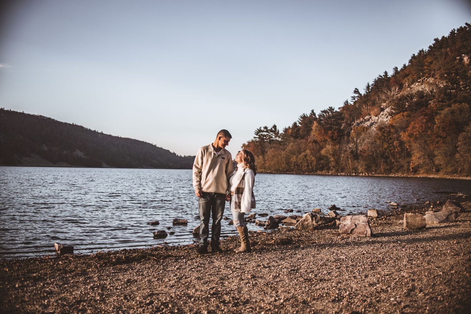 A couple stands together on a rocky lakeshore with mountains in the background at dusk.