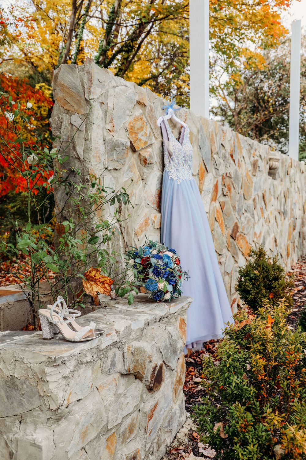 Light blue wedding dress and shoes displayed on stone wall with autumn foliage.