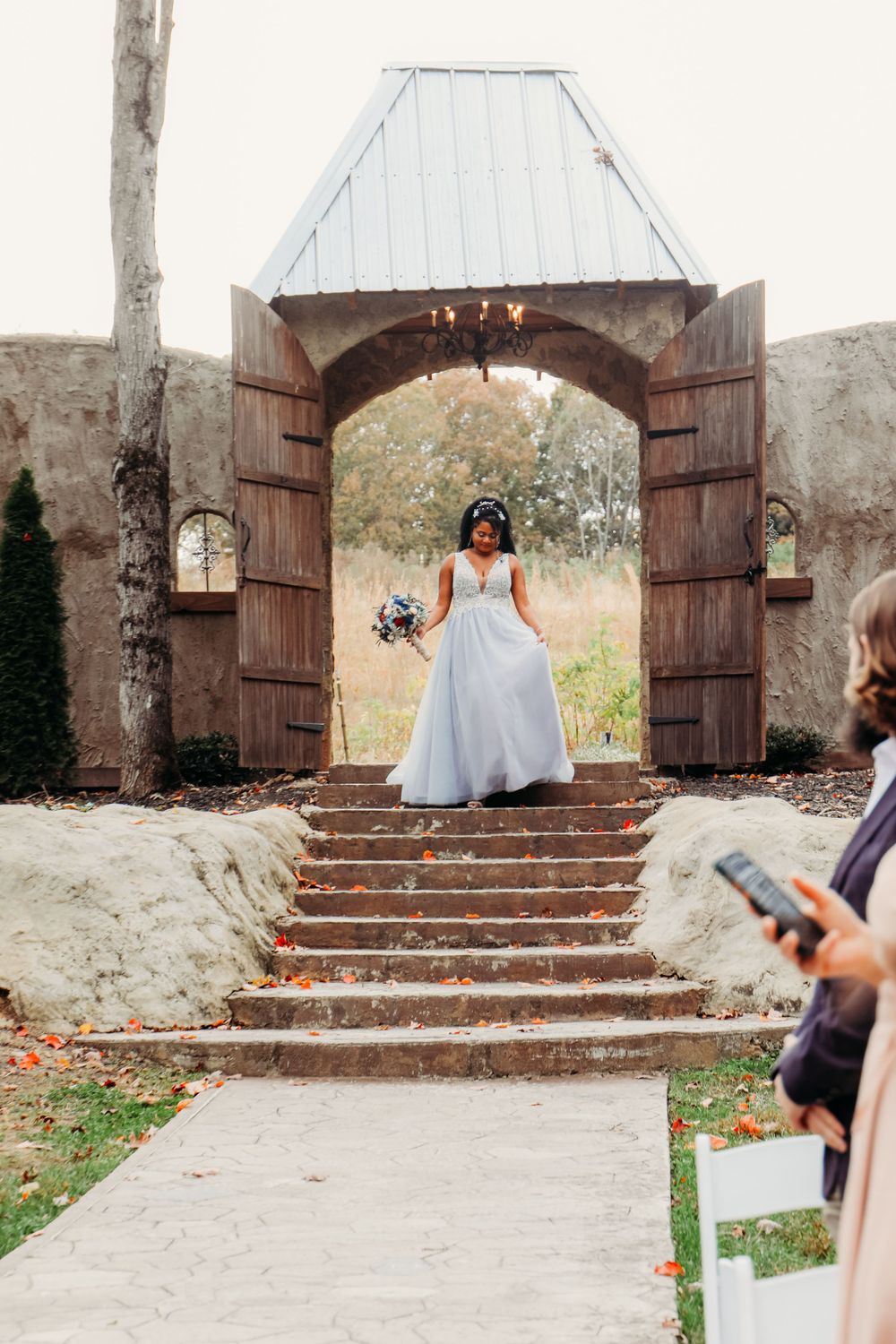 Bride in white dress stands under rustic stone archway on outdoor wedding steps.
