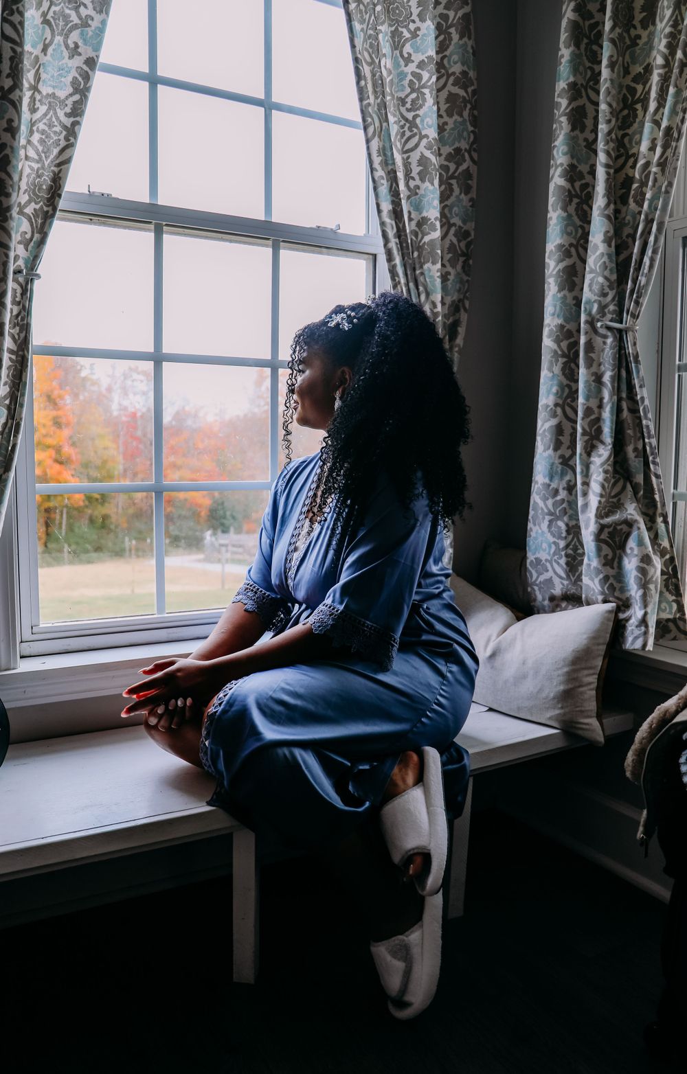 Silhouette of person sitting by window with patterned curtains and fall scenery outside.