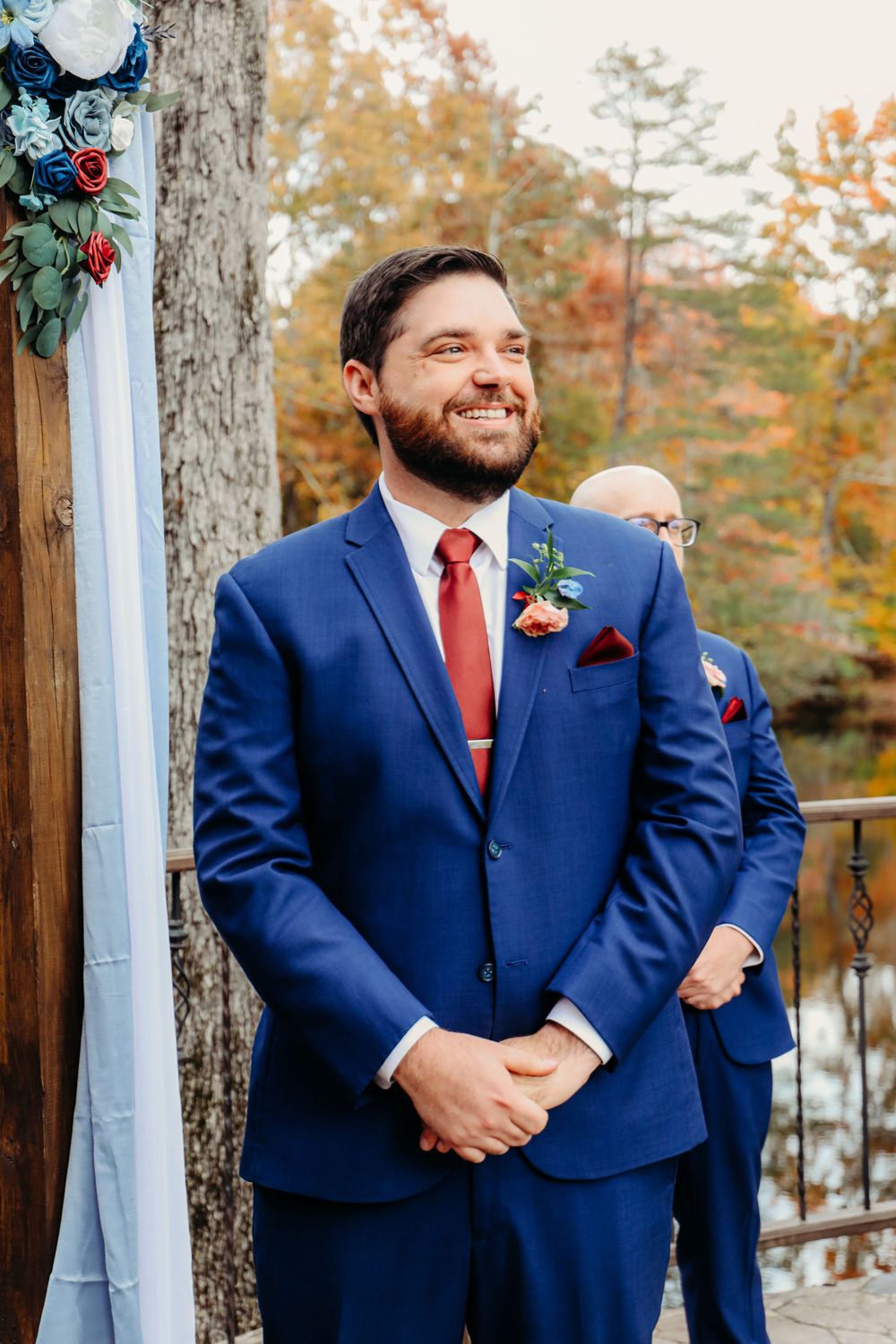 A groom in a navy blue suit and red tie smiles during an autumn wedding ceremony.