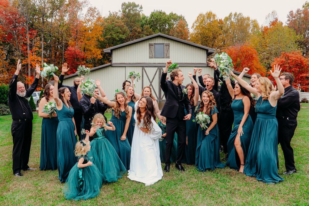 Wedding party in teal dresses and black suits pose cheerfully against autumn backdrop.
