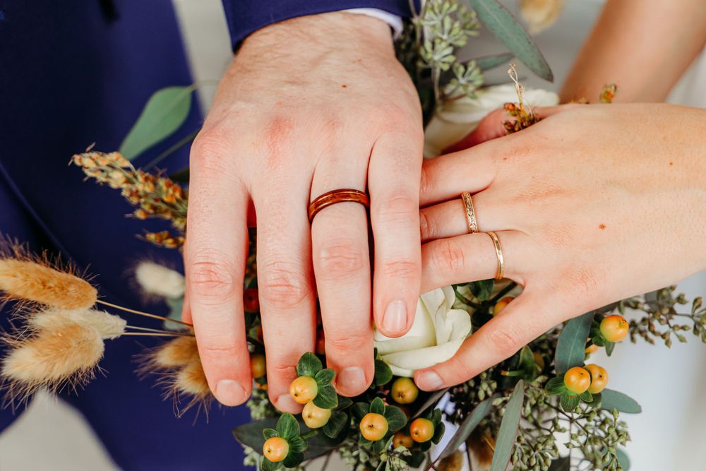 Close-up of newlywed hands with rings holding wedding bouquet.