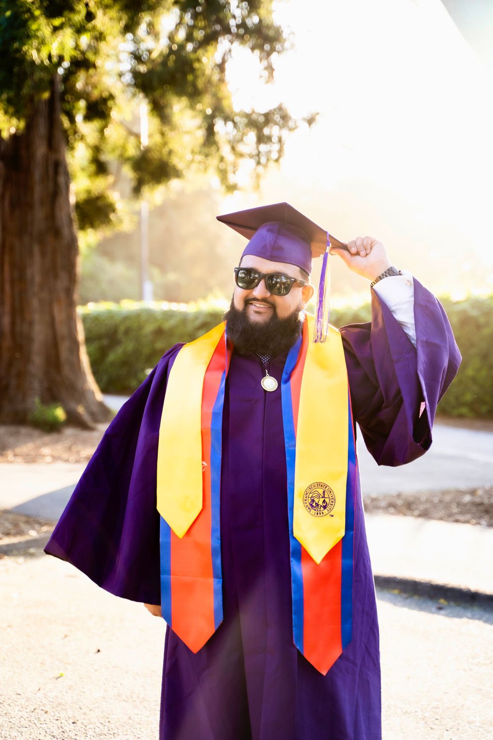 A graduate in purple cap and gown with orange and yellow stoles poses under sunlit trees during the commencement ceremony.