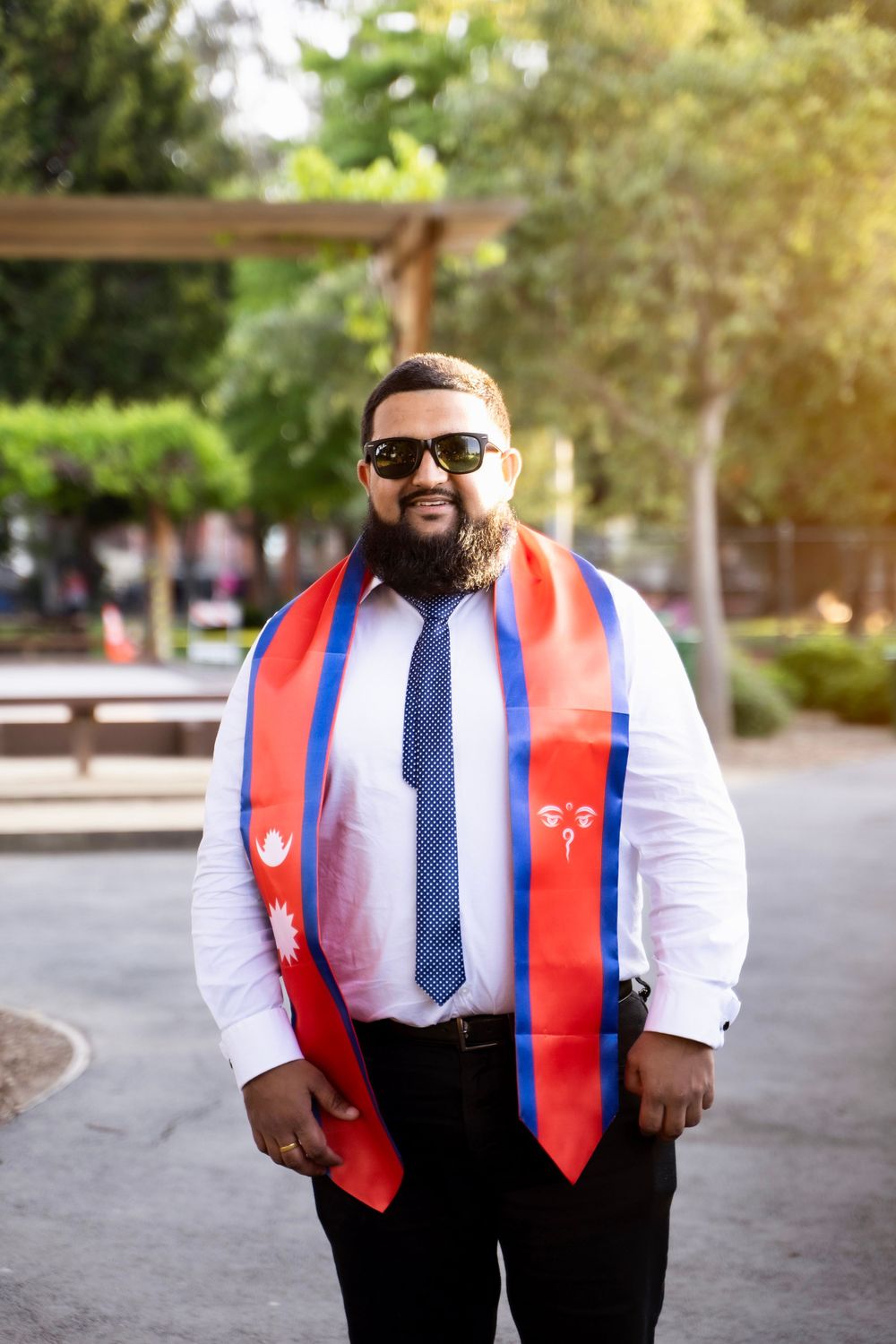 Graduate wearing red white and blue stoles with sunglasses and tie stands smiling outdoors on graduation day.