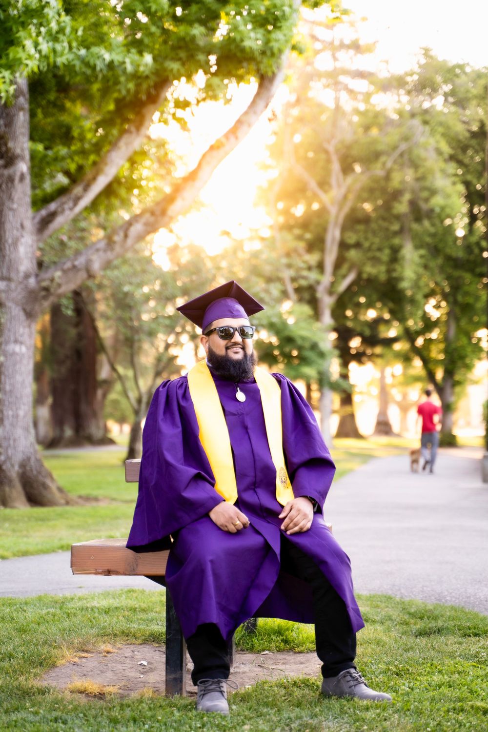 Graduate in purple cap and gown sitting on bench in park during golden hour sunset.