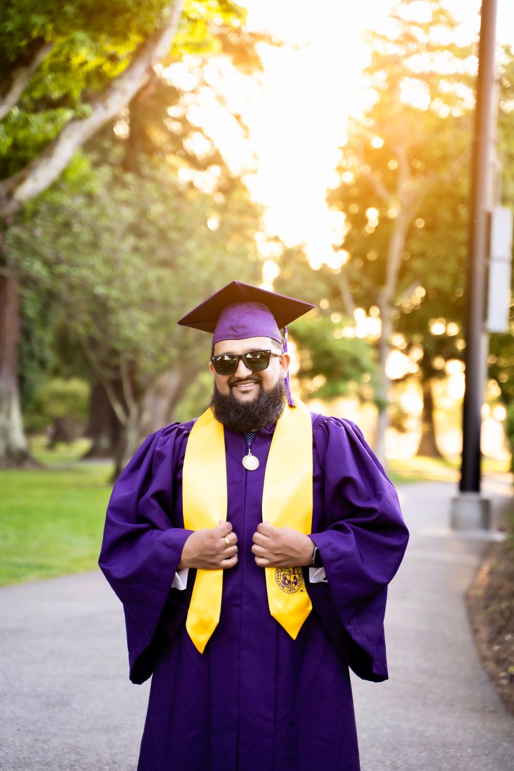 Graduate in purple cap and gown with gold stole stands smiling on campus path during golden sunset.